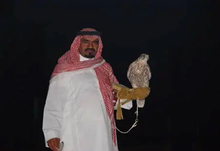 صقّار بالشماغ الأحمر والثوب الأبيض يحمل صقراً على يده في الليل
