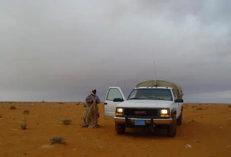 رجل واقف بجوار سيارة بيضاء مضاءة الأنوار تحت سماء ملبّدة بالغيوم